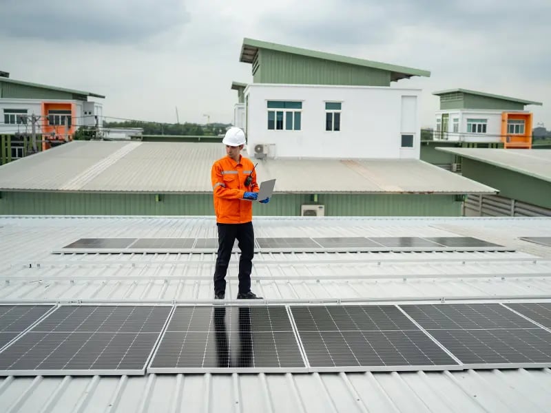 Monteur in oranger Arbeitsjacke und weißem Helm steht mit einem Laptop auf einem Trapezblechdach zwischen montierten Solarmodulen