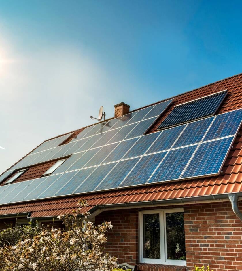 Klinkerhaus mit rotem Ziegeldach, darauf eine großflächige Photovoltaikanlage und eine Röhrenkollektor-Reihe vor blauem Himmel