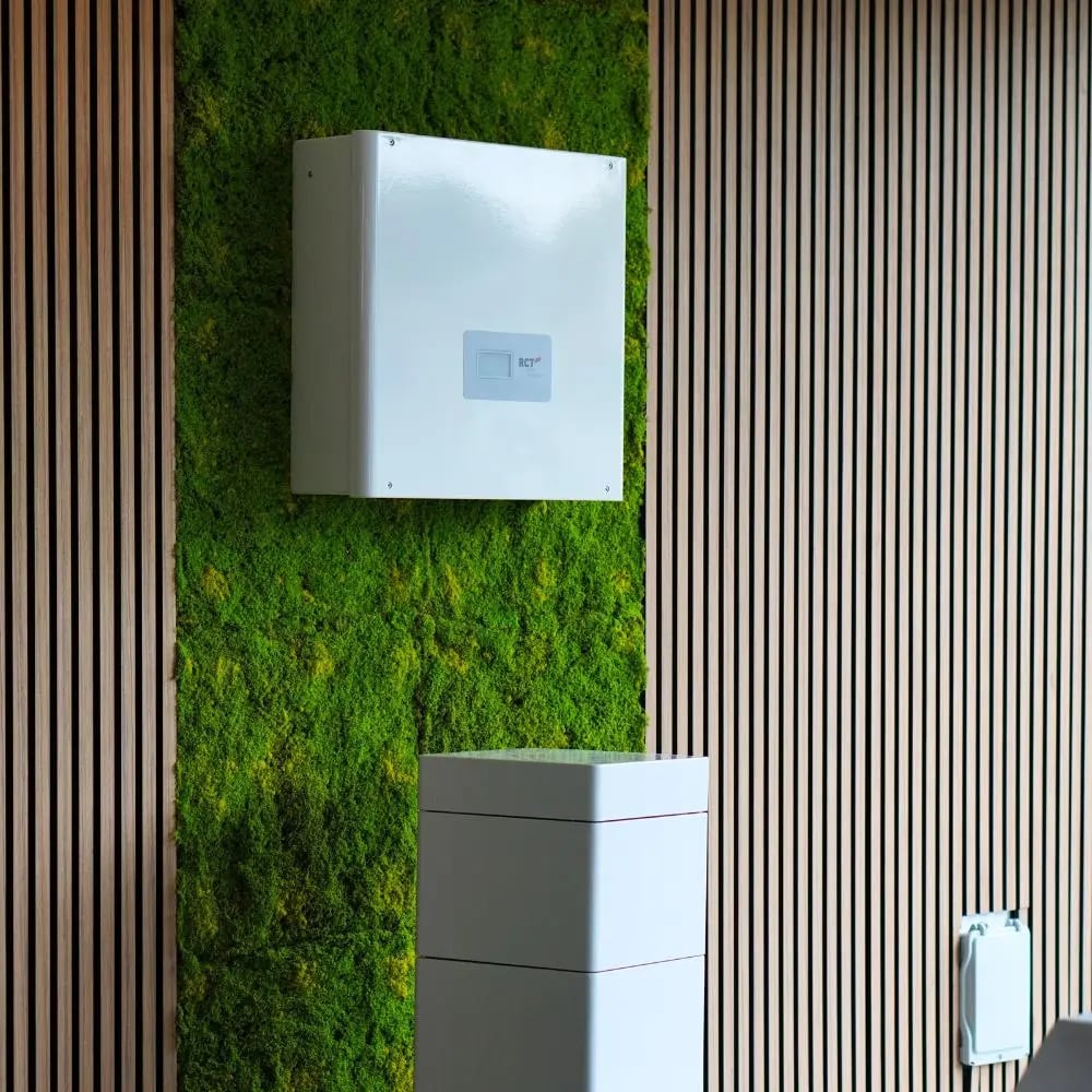 Nahaufnahme der Mooswand mit Neon-Logo zwischen Holzlamellen, davor ein schwarzes Solarmodul auf dem Boden